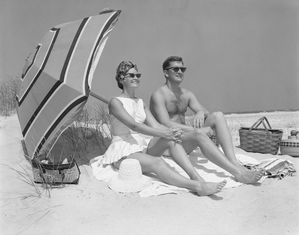 Detail of 1960s couple in sunglasses sitting on beach blanket with legs extended with umbrella by Anonymous
