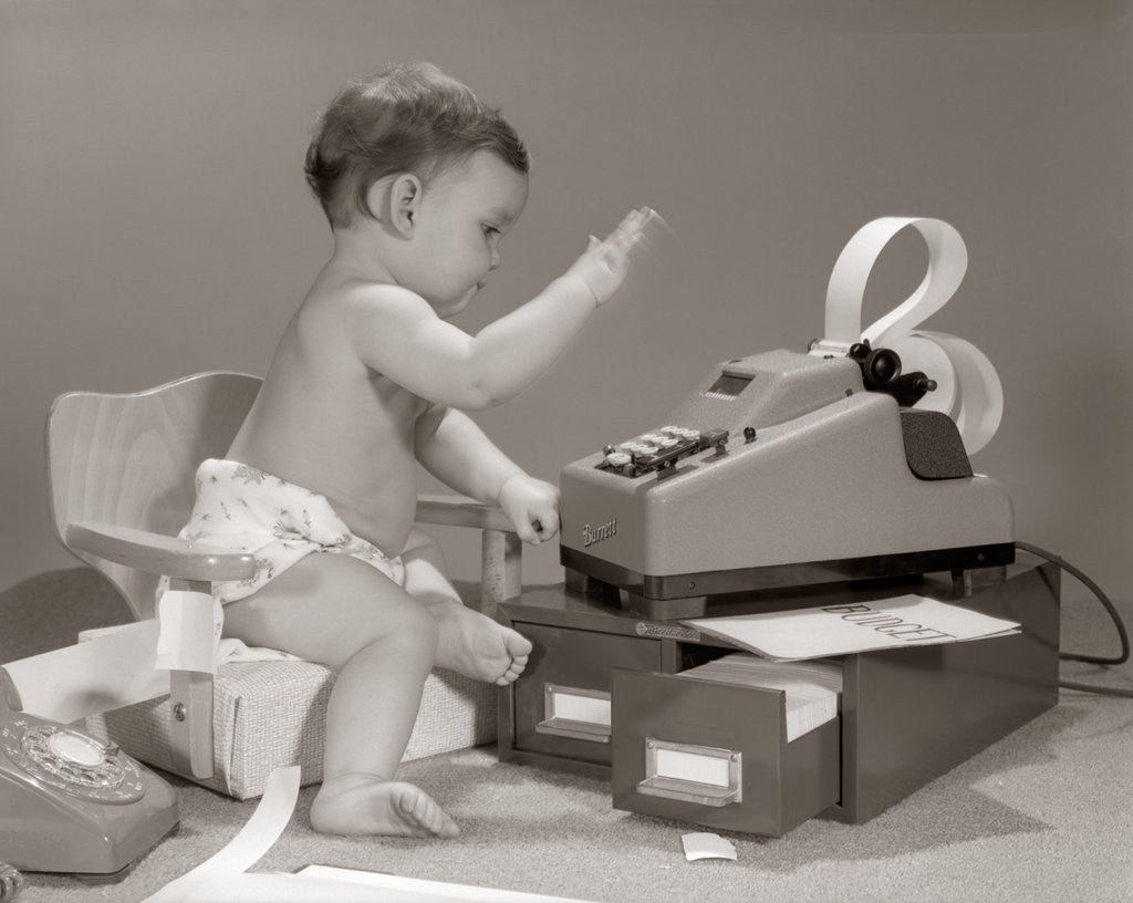 Detail of 1960s baby seated in small chair hitting keys on office adding machine on top of small file drawers by Anonymous