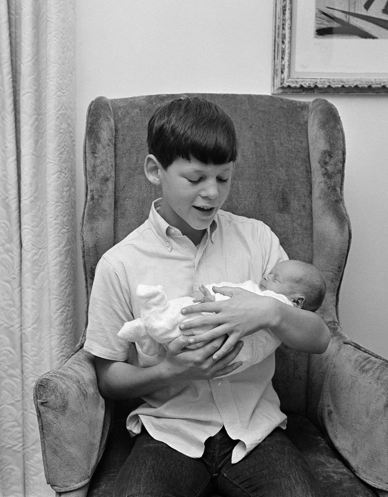 Detail of 1960s pre-teen boy sitting in armchair holding infant newborn baby sibling by Anonymous