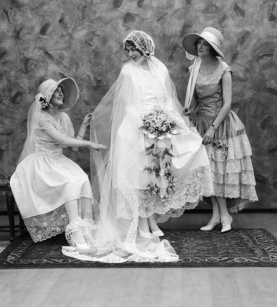 Detail of 1900 1910s bride with one bridesmaid on either side helping fix her wedding dress by Anonymous