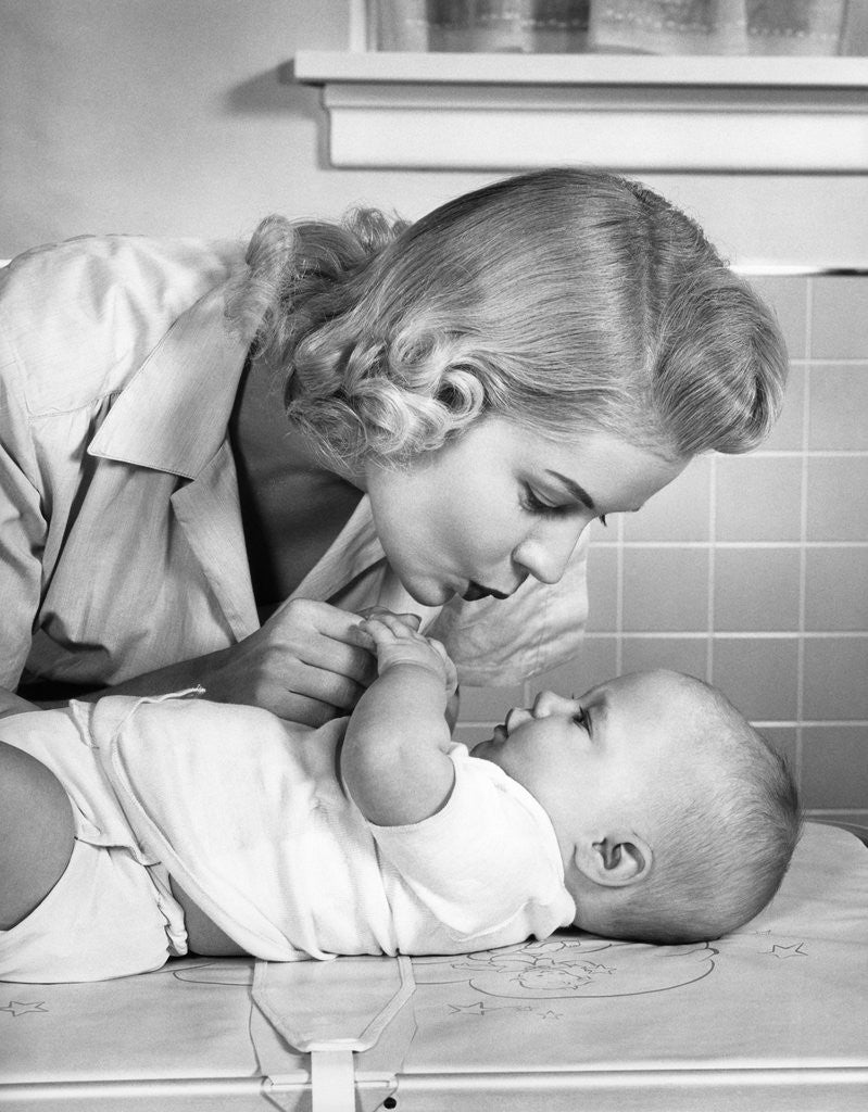 Detail of 1950s mother woman bending over baby lying on layette holding hands talking by Anonymous
