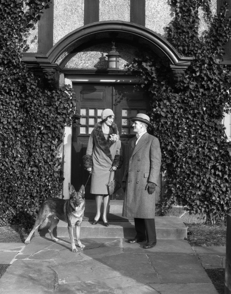 1920s couple wearing coat hat gloves on steps ivy covered building with german shepherd dog on a leash by Corbis