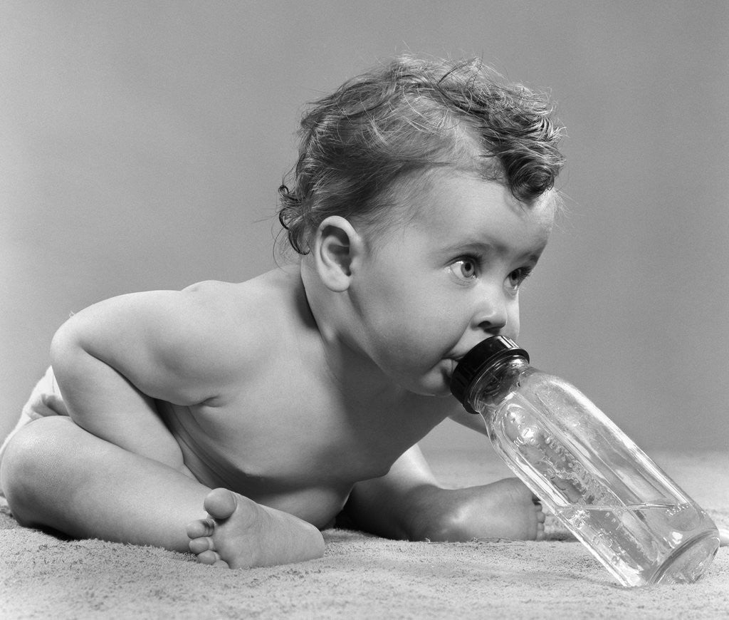 Detail of 1950s baby sitting leaning forward drinking from bottle studio indoor by Anonymous