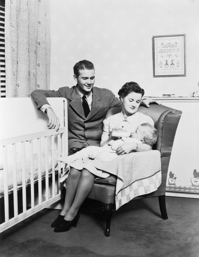 Detail of 1930s 1940s mother sitting in chair in nursery baby 11 months on lap drinking from bottle father looking on leaning on crib by Anonymous