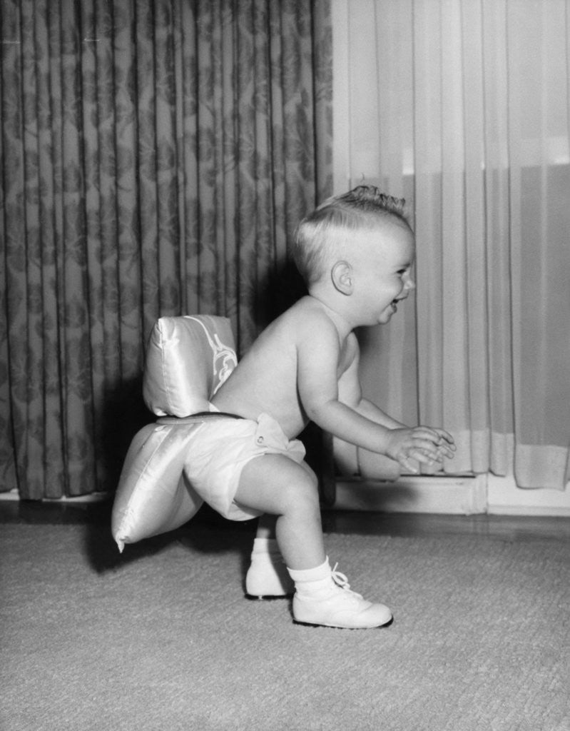 1950s laughing baby in diaper and shoes learning to walk with a pillow tied to his rear end by Corbis