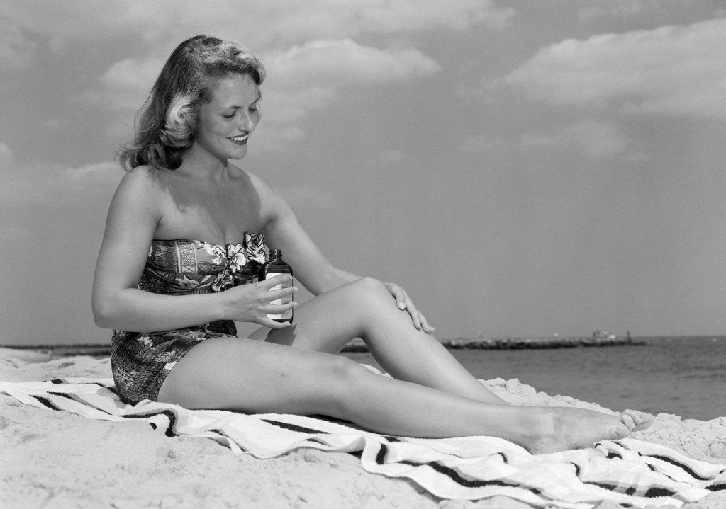 Detail of 1950s woman in strapless one-piece bathing suit seated on beach towel putting on suntan lotion by Anonymous