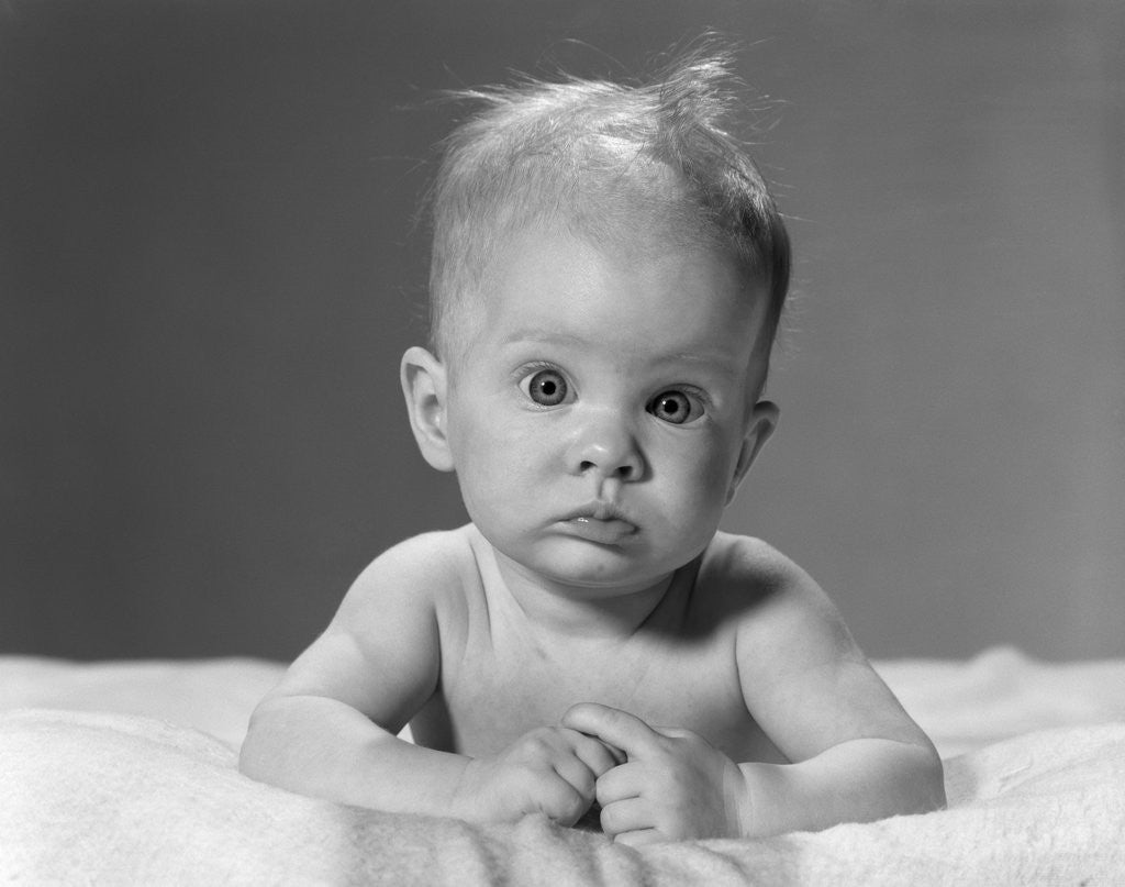 Detail of 1960s portrait baby lying on stomach with messy hair and bulging eyes looking at camera by Corbis