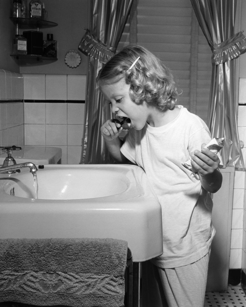 Detail of 1950s little blond girl in pajamas standing at bathroom sink brushing her teeth toothpaste and toothbrush in hand by Anonymous