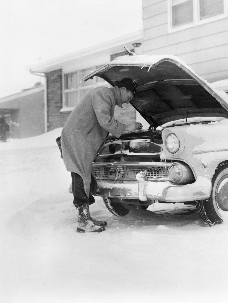 Detail of 1950s man winter driveway trying to start fix car hood up by Anonymous