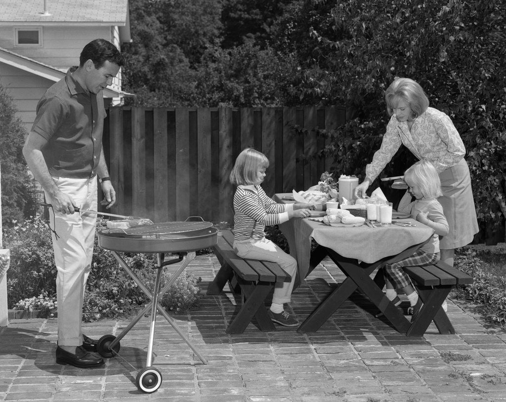 Detail of 1960s man on patio grilling steak with 2 daughters seated at picnic table & wife standing serving food by Anonymous