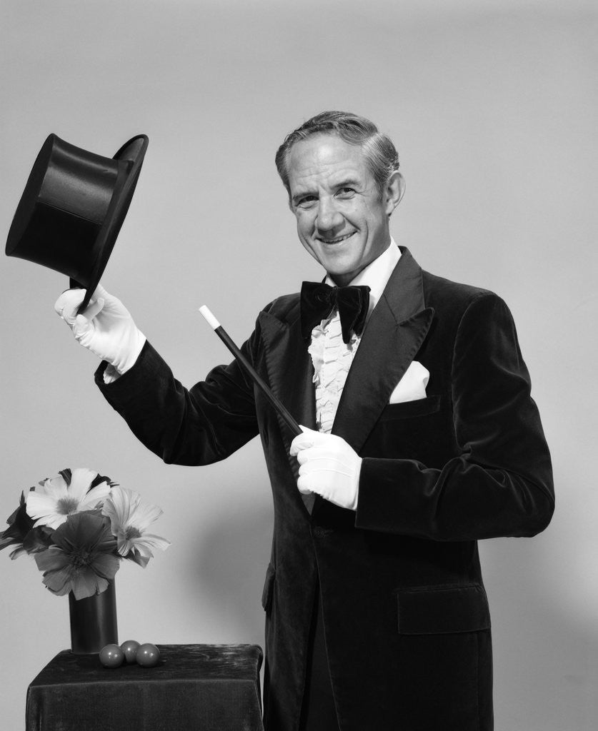 Detail of 1970s smiling man magician wearing velvet tuxedo white gloves pointing magic wand at top hat looking at camera by Corbis