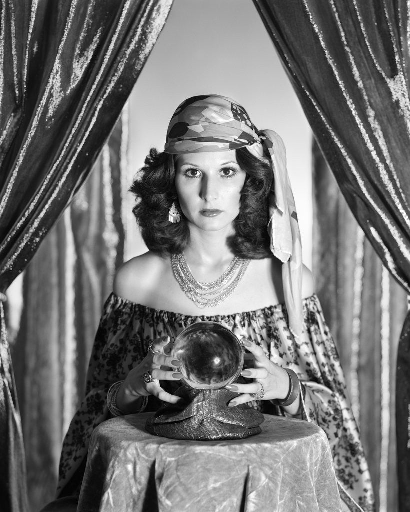 Detail of 1970s gypsy woman between sparkly curtains with hands around crystal ball staring into camera by Anonymous
