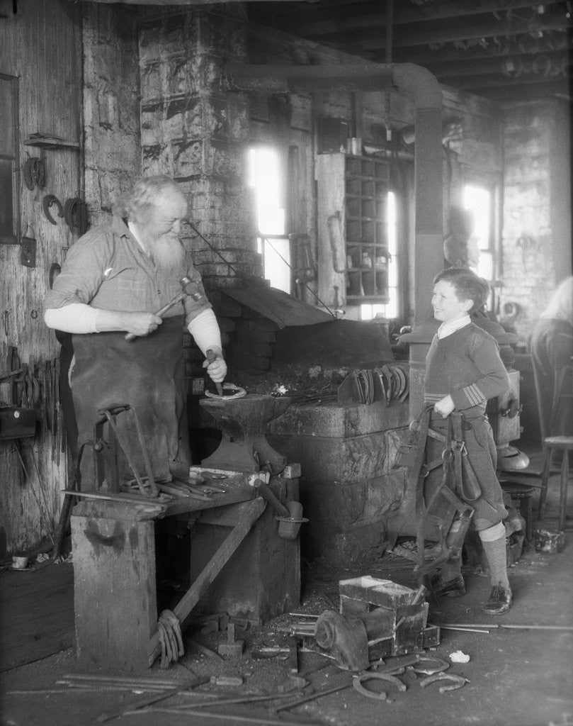 Detail of 1930s elderly blacksmith with hammer at anvil as young boy holding harness looks on smiling by Corbis