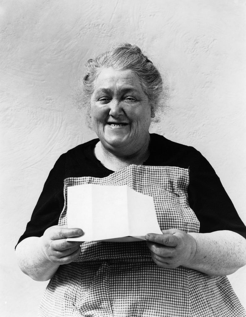 Detail of 1930s 1940s elderly character woman smiling reading letter wearing apron looking at camera by Corbis
