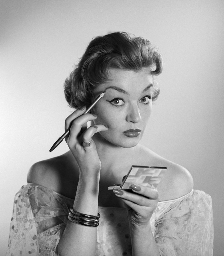 Detail of 1950s 1960s woman looking at camera applying makeup eye brow pencil holding compact mirror by Anonymous