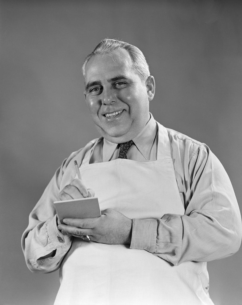 Detail of 1940s man wearing white apron writing order on pad with pencil looking at camera by Anonymous