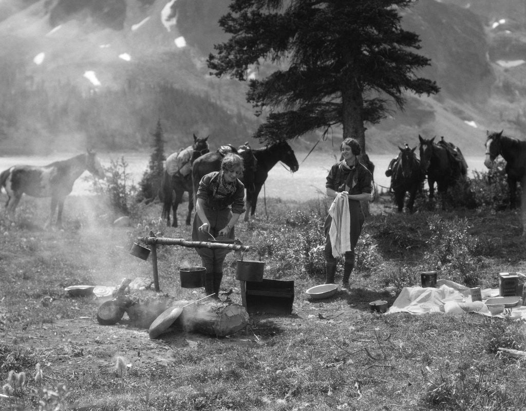 Detail of 1920s 1930s two women at campsite woman cooking preparing food over campfire horses with riding gear in background by Corbis