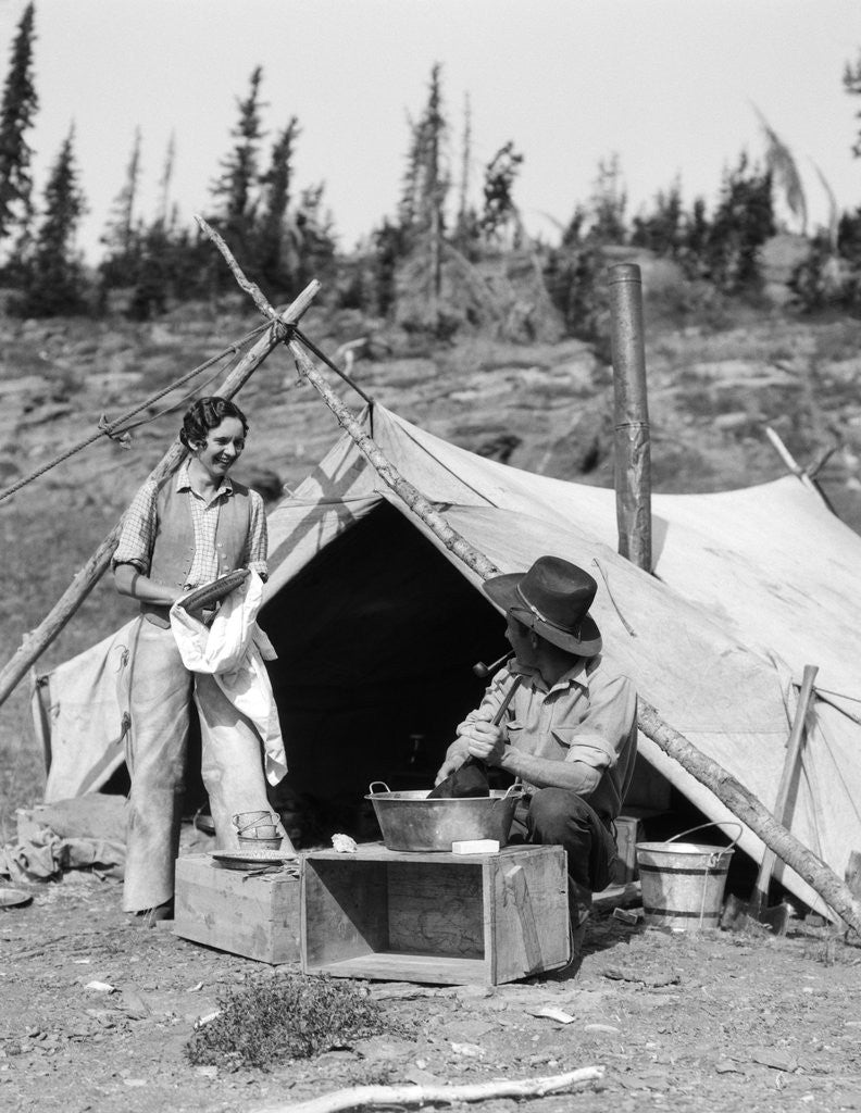 Detail of 1930s smiling talking couple working by rustic western campsite tent man in cowboy hat smoking pipe washing skillet woman drying dishes by Corbis