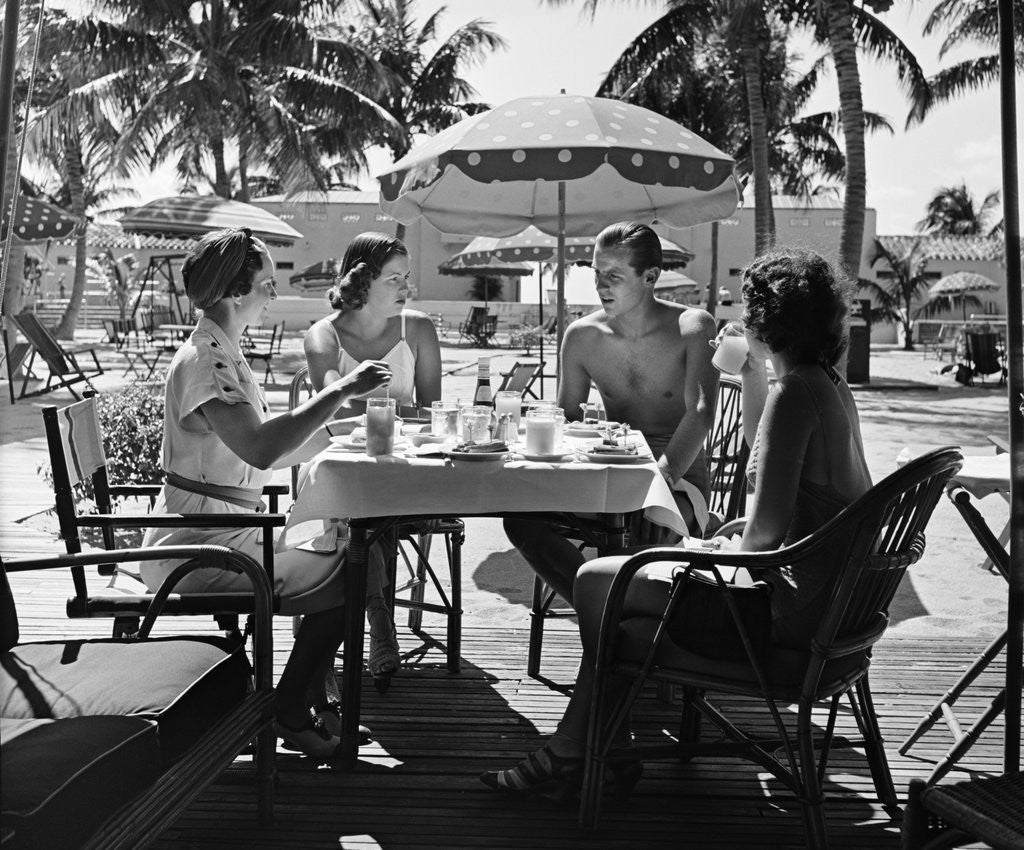 Detail of 1930s three women and one man sitting at tropical pool side table talking together by Anonymous