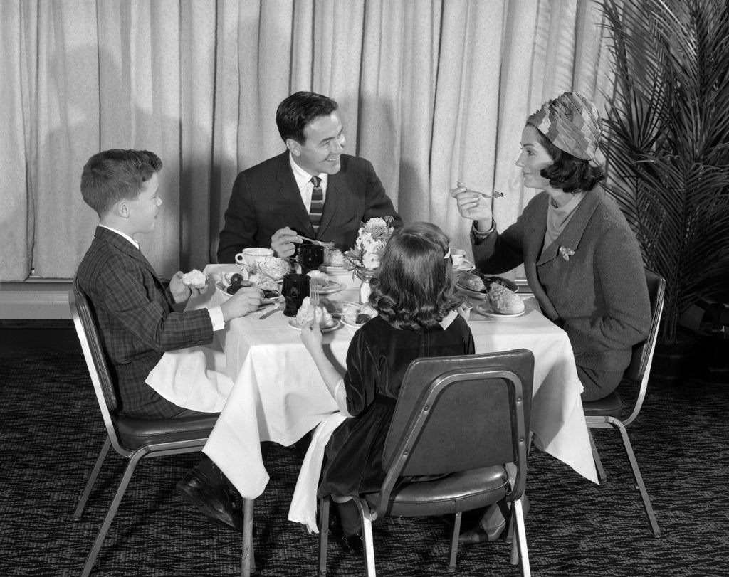 Detail of 1960s smiling happy family of four eating meal in restaurant dad talking to mom who is wearing turban style hat by Anonymous