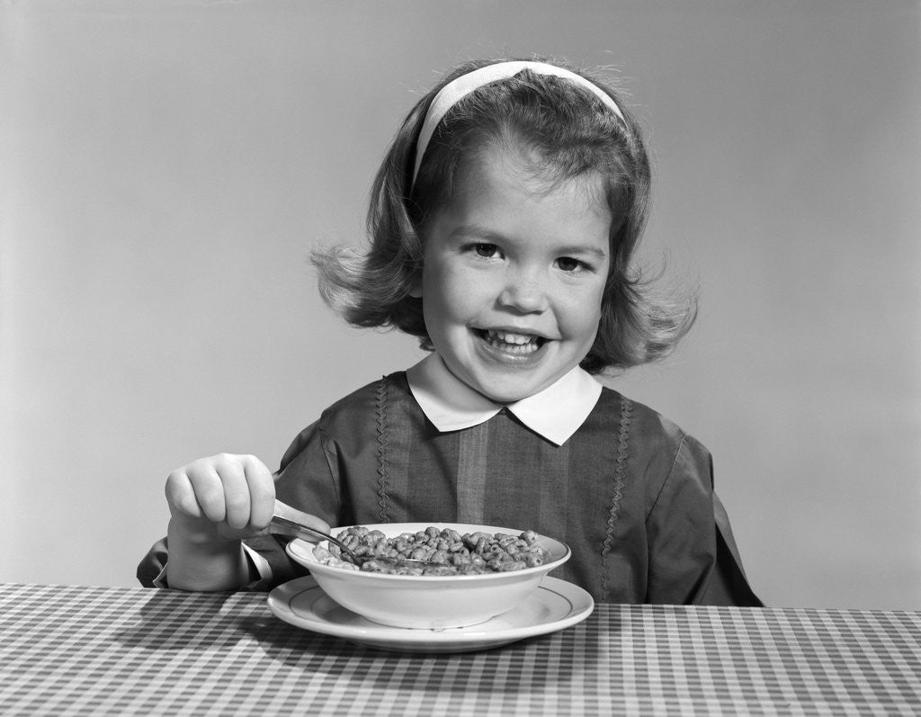 Detail of 1950s 1960s smiling little girl eating a bowl of breakfast cereal looking at camera by Anonymous