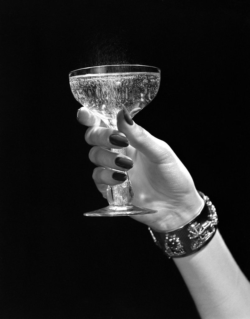 Detail of 1930s 1940s 1950s woman hand ornate metal bracelet holding up new year toast glass of champagne against black background by Anonymous
