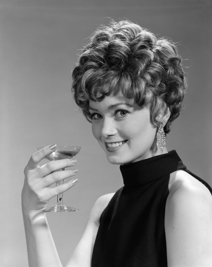 Detail of 1960s glamorous woman with curly hair and dangling earrings smiling holding a champagne glass looking at camera by Anonymous