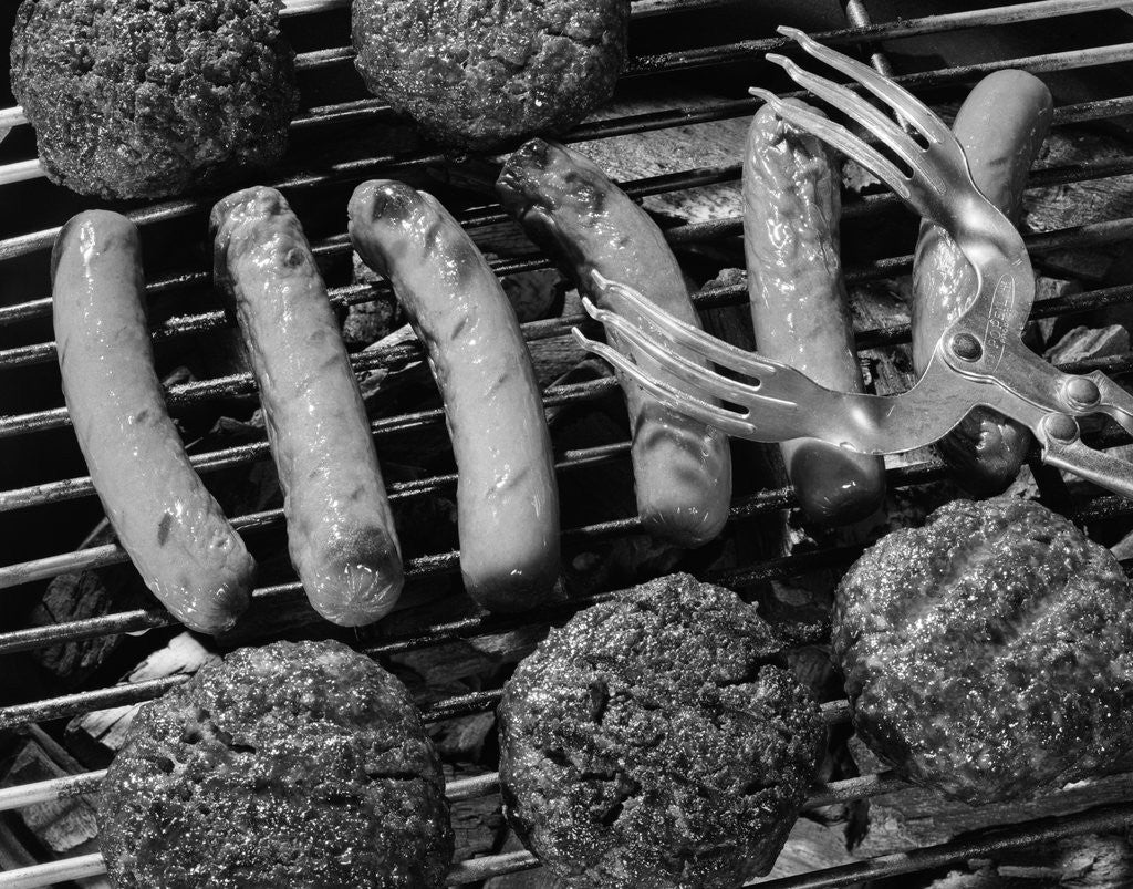 Detail of 1950s close-up of grill filled with hot dogs & hamburgers & tongs reaching down to turn them over by Anonymous