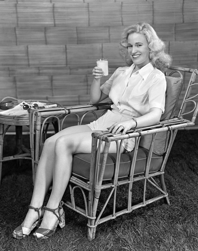Detail of 1940s blonde woman in bamboo chair holding up glass of milk smiling wearing white shorts looking at camera by Anonymous