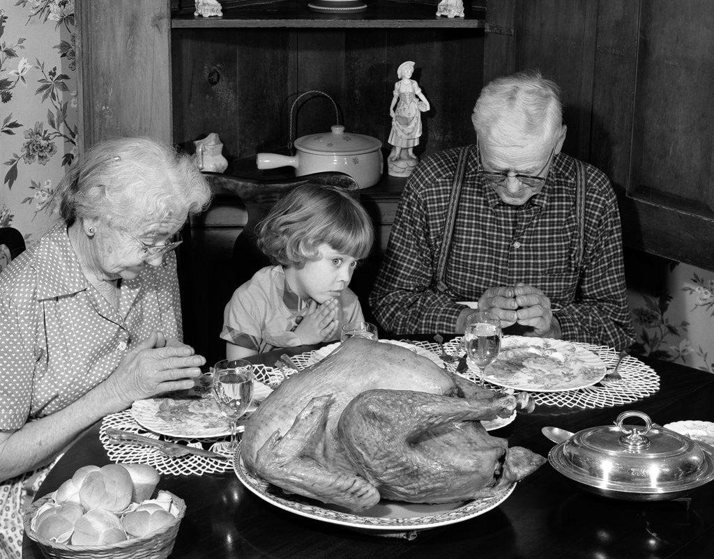 Detail of 1950s 1960s little girl look at turkey sitting between grandmother grandfather saying grace at table by Anonymous