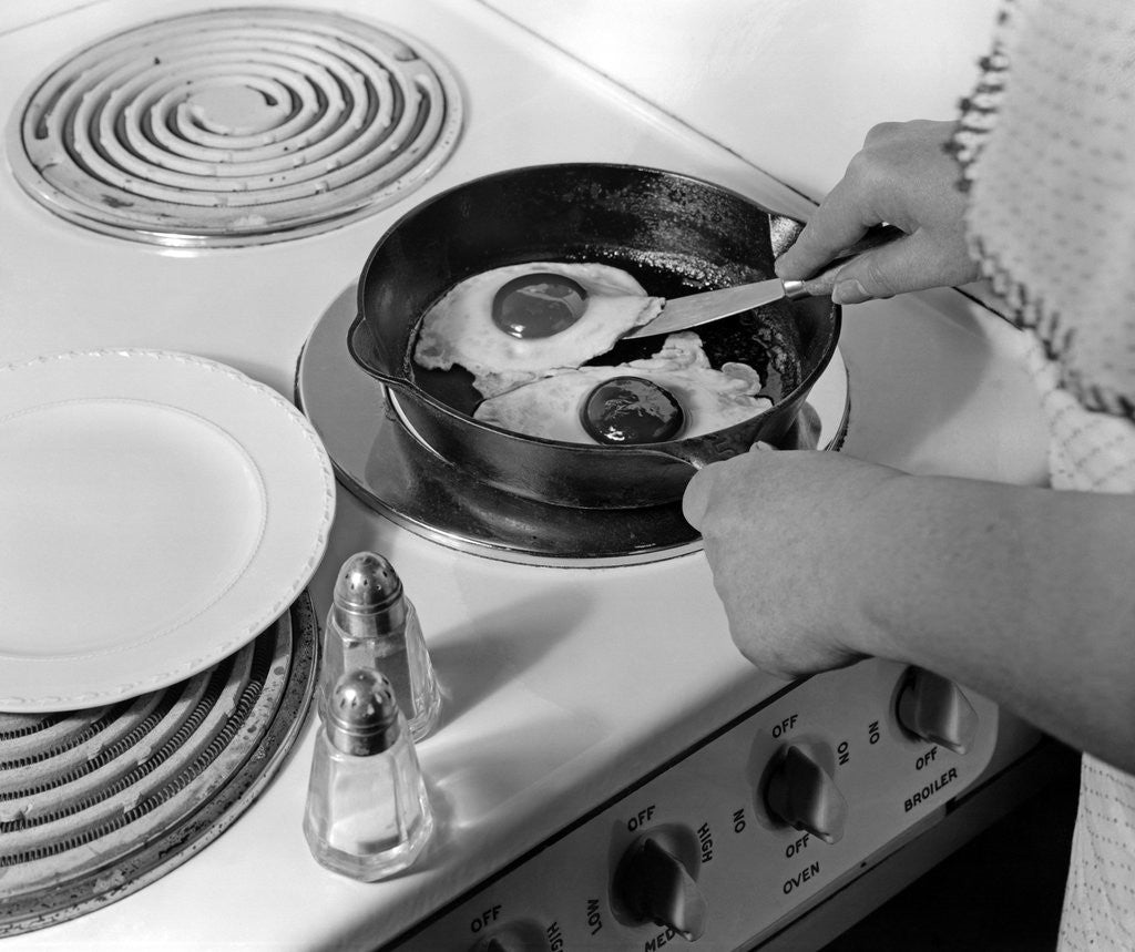 Detail of 1940s 1950s woman hands frying eggs in iron skillet on electric stove salt and pepper shakers by Anonymous