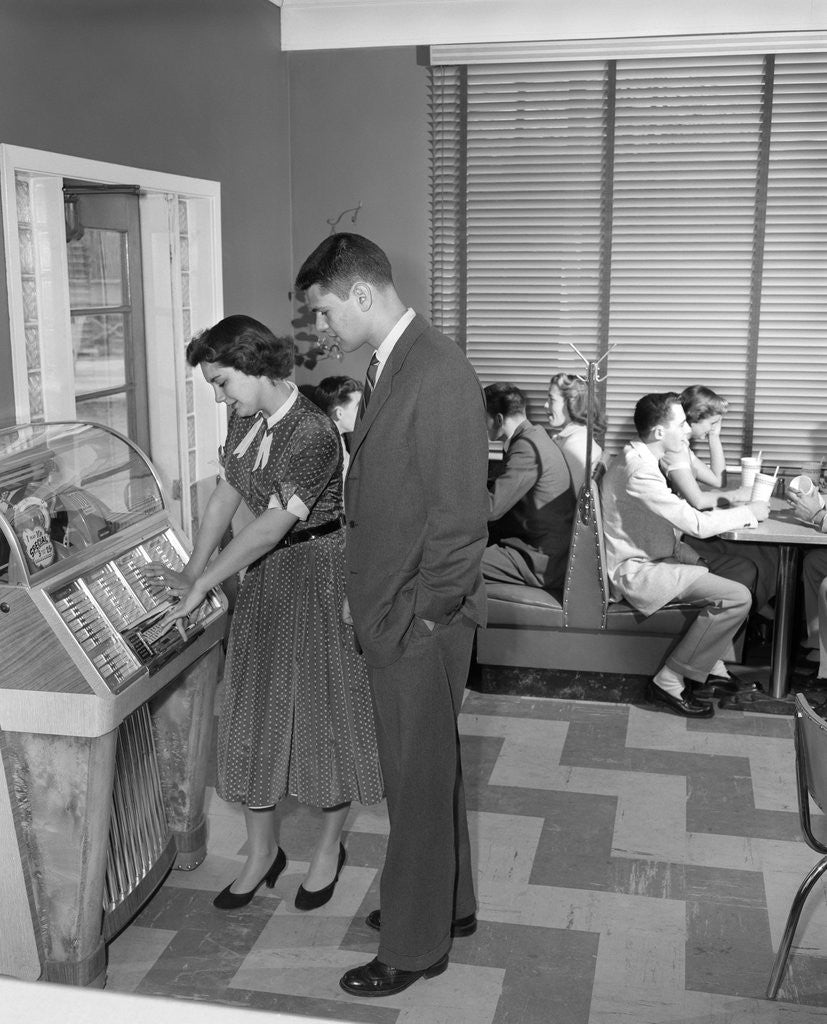 Detail of 1950s teen couple playing juke box in malt shop with other teens in booths by Anonymous