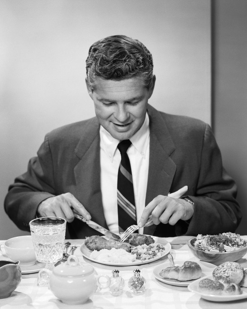 Detail of 1950s smiling man in suit and tie sitting at table holding knife and fork eating dinner by Anonymous