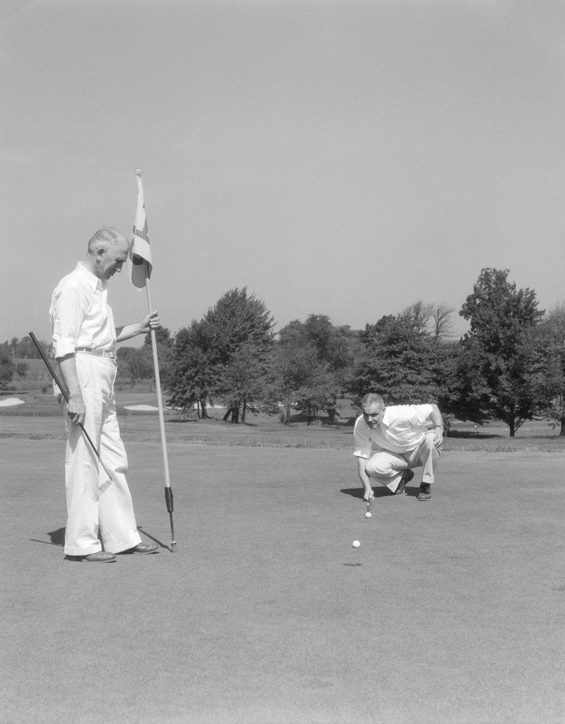 Detail of 1930s 1940s elderly men on golf green one holding flag the other kneels lining up his putt by Anonymous