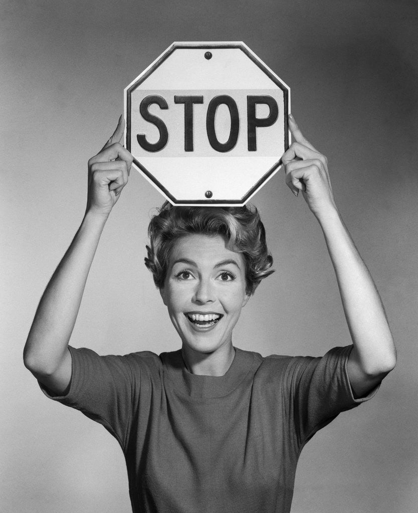 Detail of 1950s 1960s smiling woman holding stop sign above her head looking at camera by Anonymous