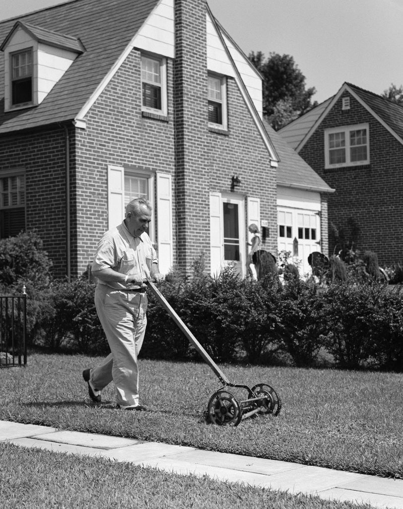 Detail of 1940s 1950s elderly overweight man pushing lawn mower in front of brick house by Anonymous
