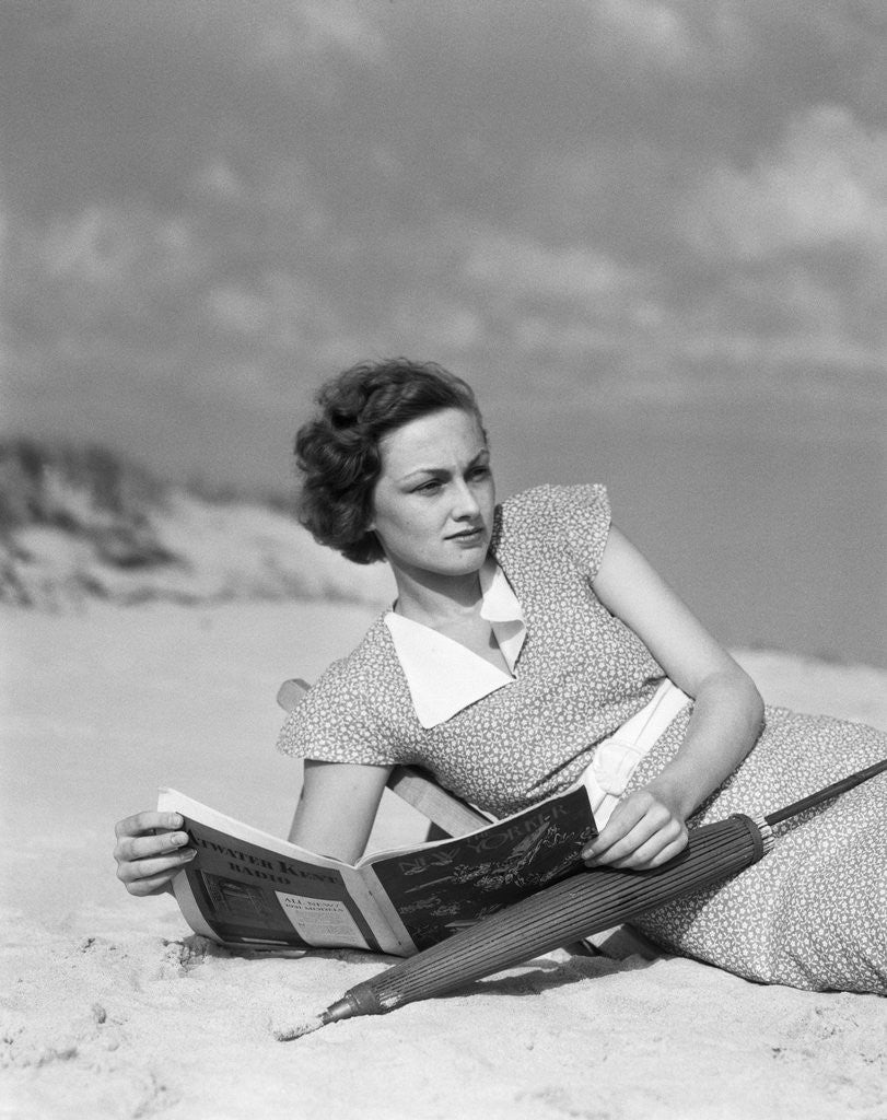 Detail of 1930s woman leaning in wooden chair on beach wearing printed dress holding magazine looking thoughtful by Anonymous