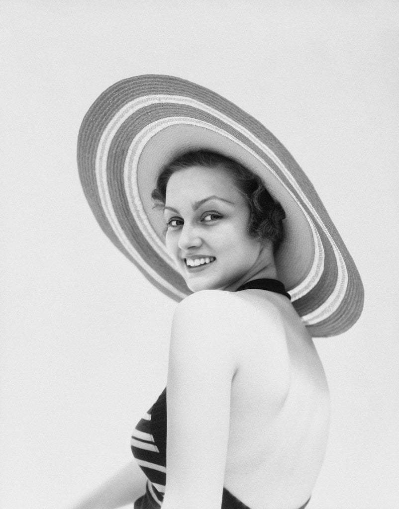Detail of 1930s woman wearing striped halter top and very wide brim straw hat looking at camera over shoulder by Anonymous