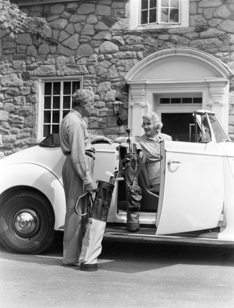 Detail of 1940s senior retired couple loading golf clubs into white convertible car in front stone house by Anonymous