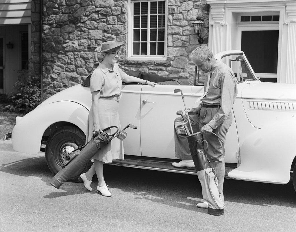 Detail of 1930s 1940s senior couple putting golf clubs in convertible car by stone house by Corbis