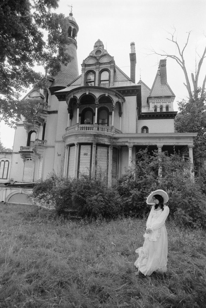 Detail of 1970s woman in victorian costume standing on front lawn of large abandoned haunted victorian home by Anonymous