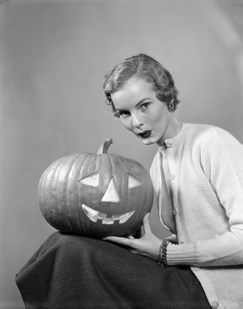 Detail of 1950s woman wearing sweater holding carved pumpkin jack-o-lantern on her lap by Anonymous