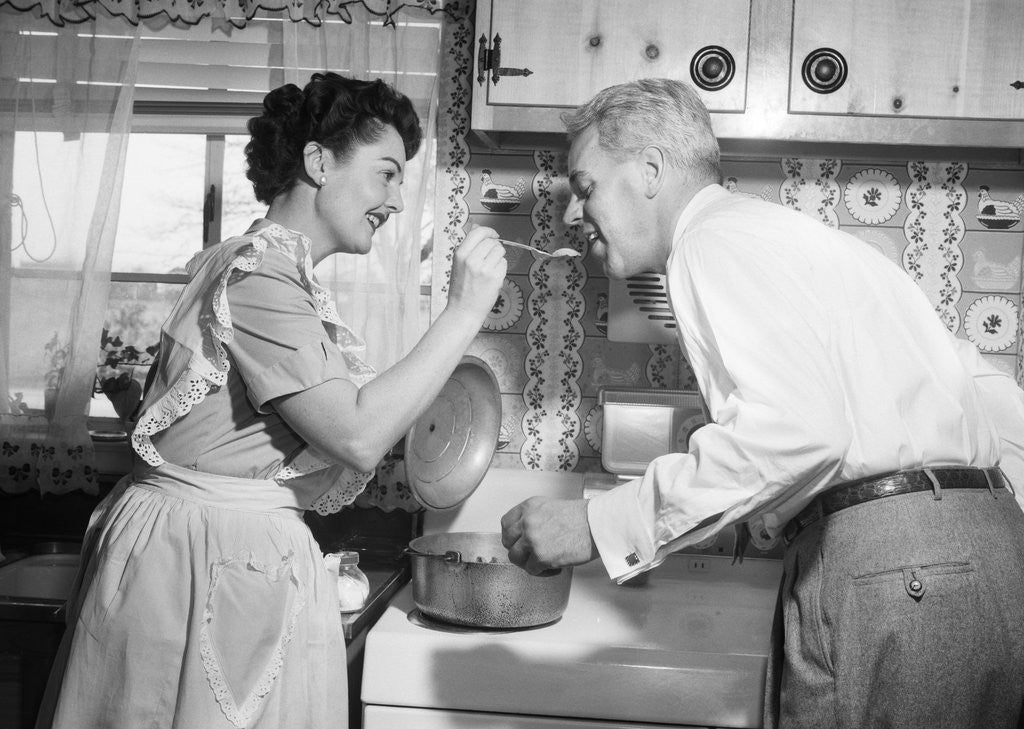 Detail of 1950s housewife in kitchen having husband taste food on stove by Anonymous