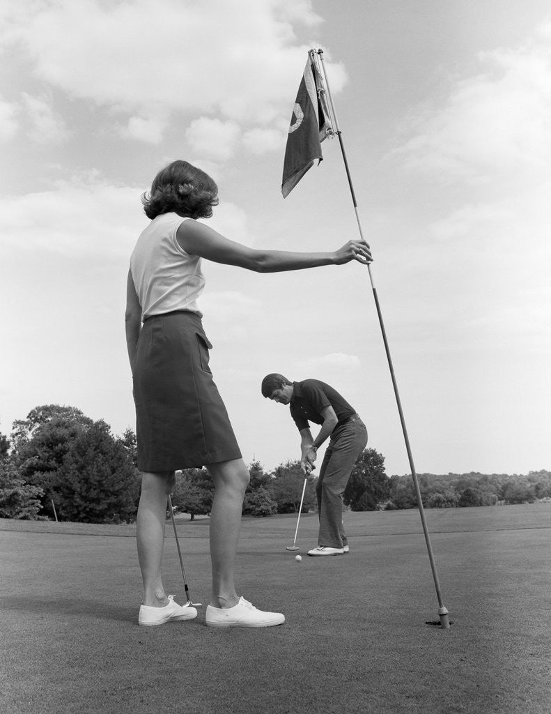 Detail of 1970s couple playing golf man putting woman holding flag by Corbis