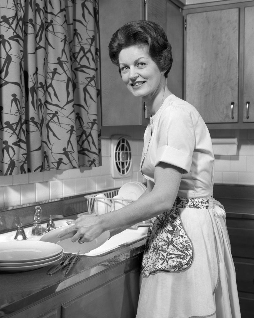 Detail of 1960s woman housewife washing dishes in kitchen sink looking at camera by Anonymous