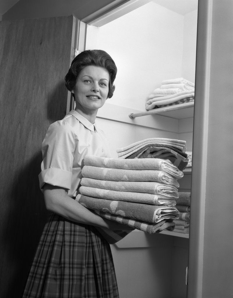 Detail of 1950s 1960s housewife holding laundry folded towels by linen closet looking at camera by Anonymous