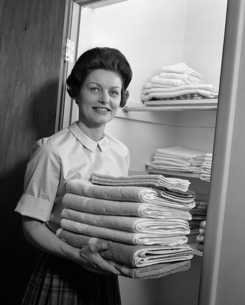Detail of 1960s woman holding laundry folded towels by linen closet looking at camera by Anonymous