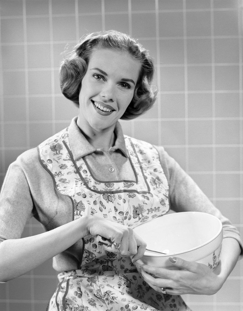 Detail of 1950s smiling woman wearing apron in kitchen holding mixing bowl and spoon looking at camera by Anonymous
