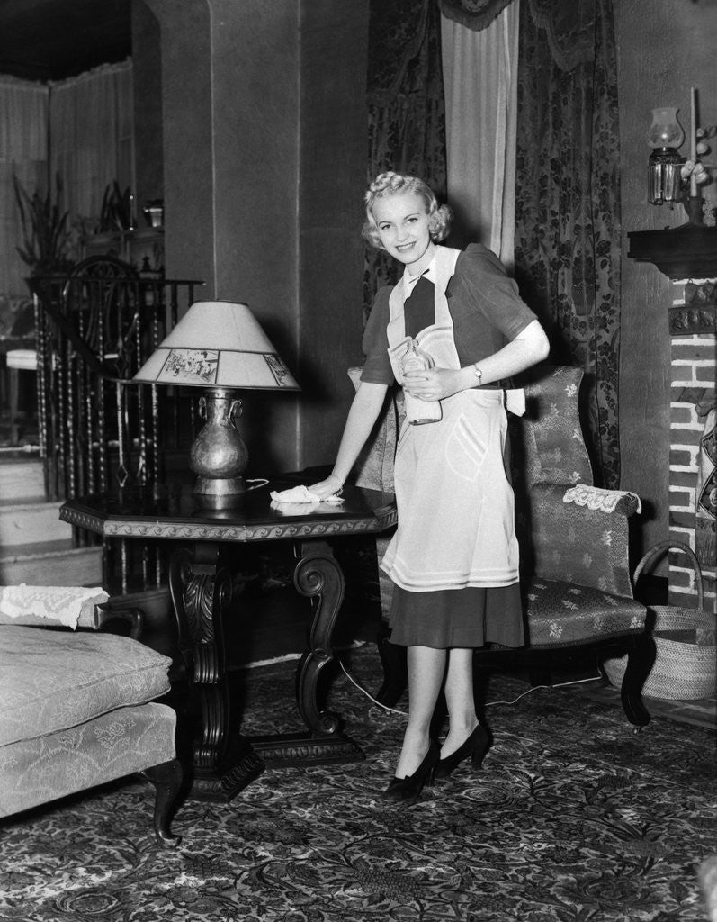 Detail of 1940s blonde woman housewife maid wearing apron cleaning polishing wooden end table in ornate living room by Anonymous
