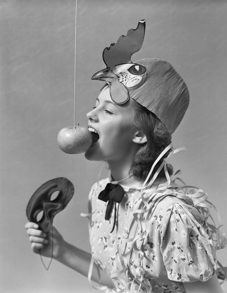Detail of 1930s 1940s girl bobbing for apple dangling on a string wearing party hat and holding eye mask by Corbis